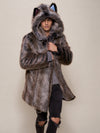 A model wearing a long, plush faux fur coat with a hood featuring cat ears and a striped pattern in shades of gray, brown, and black, posed against a solid beige studio background.