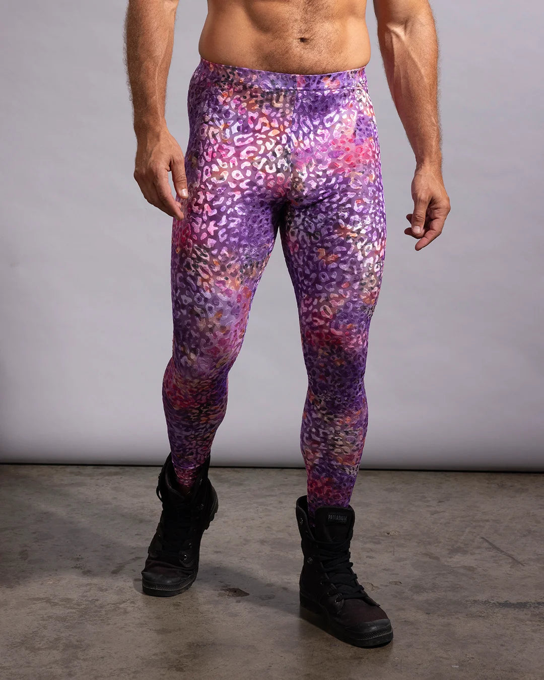 A man wearing high-waisted colorful leggings with a purple, pink, and orange leopard-like abstract pattern, paired with black boots, standing against a gray studio background.