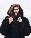 A Man wearing SpiritHoods Black Wolf Hooded Faux Fur Coat in the middle of wind and snow holding the hood with his hands looking straight into the camera. SpiritHoods 100% Fake Fur Coats and Faux Fur Jackets