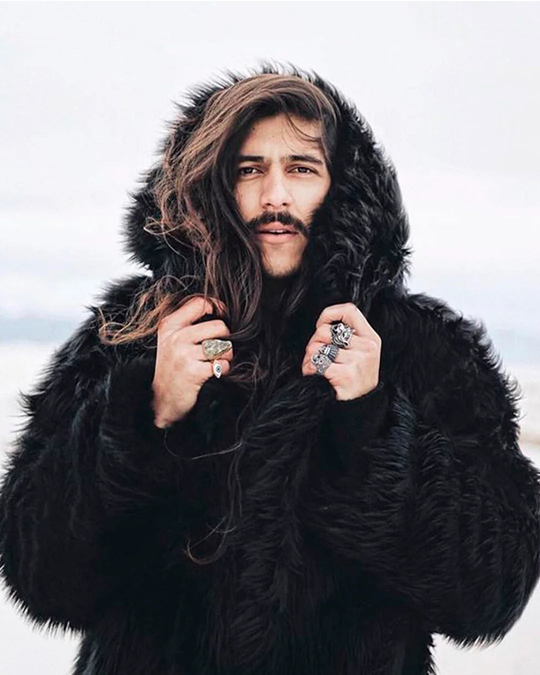A Man wearing SpiritHoods Black Wolf Hooded Faux Fur Coat in the middle of wind and snow holding the hood with his hands looking straight into the camera. SpiritHoods 100% Fake Fur Coats and Faux Fur Jackets