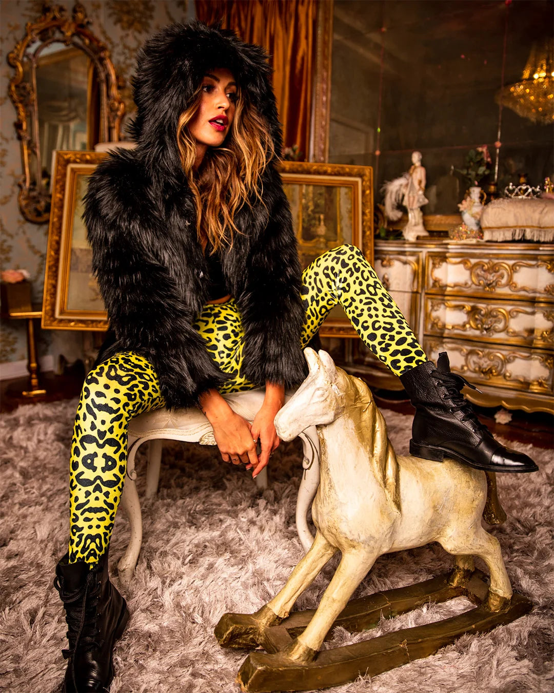 Woman sitting on a chair wearing SpiritHoods Best Selling Hooded Black Wolf Faux Fur Coat, with neon yellow leopard print leggings, as she stares to the side of the room in a playful way