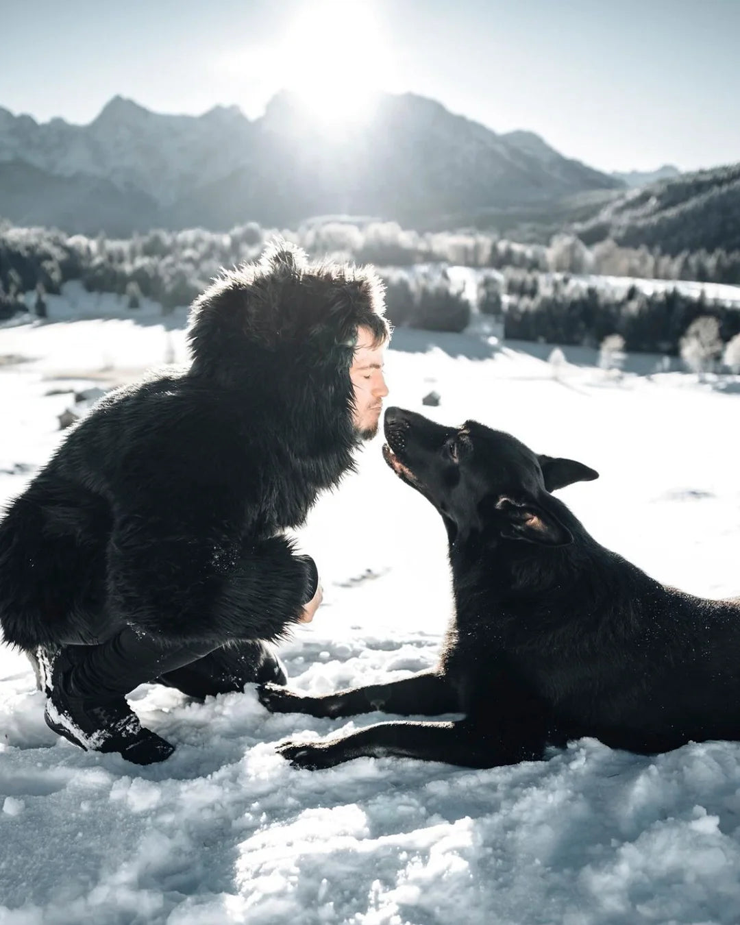 A Man wearing SpiritHoods Black Wolf Classic Faux Fur Coat with the hood up,showing wolf ears on top, leaning down in the snow, giving his all black dog a kiss in the snow with mountains in the background
