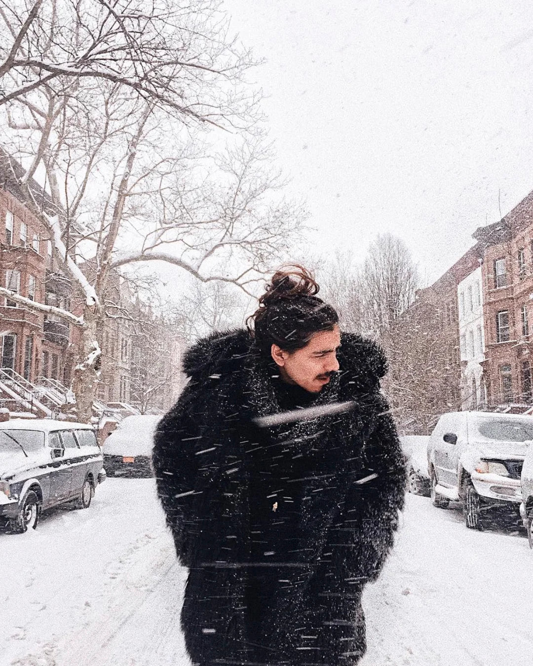 A Man braving a winter snow storm, walking down a new york city street with the wind and snow as he wears SpiritHoods Black Wolf Faux Fur Coat with his hands in the pockets to stay warm!