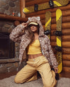 Woman wearing spirithoods Hooded Puffer Jacket in tan leopard print, while posing in a cabin with animal print skis on the walls. The faux fur puffer jacket in leopard print has the hood up, showing SpiritHoods classic ears on the top of the hood. SpiritHoods Puffer Jackets are made for the snow and always 100% vegan. 