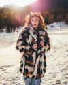 Woman wearing a patterned patchwork style plush faux fur coat standing in a snowy landscape with mountains in the background. SpiritHoods Collector Edition Manx Cat brown, black, and white fake fur jacket with an oversized collar