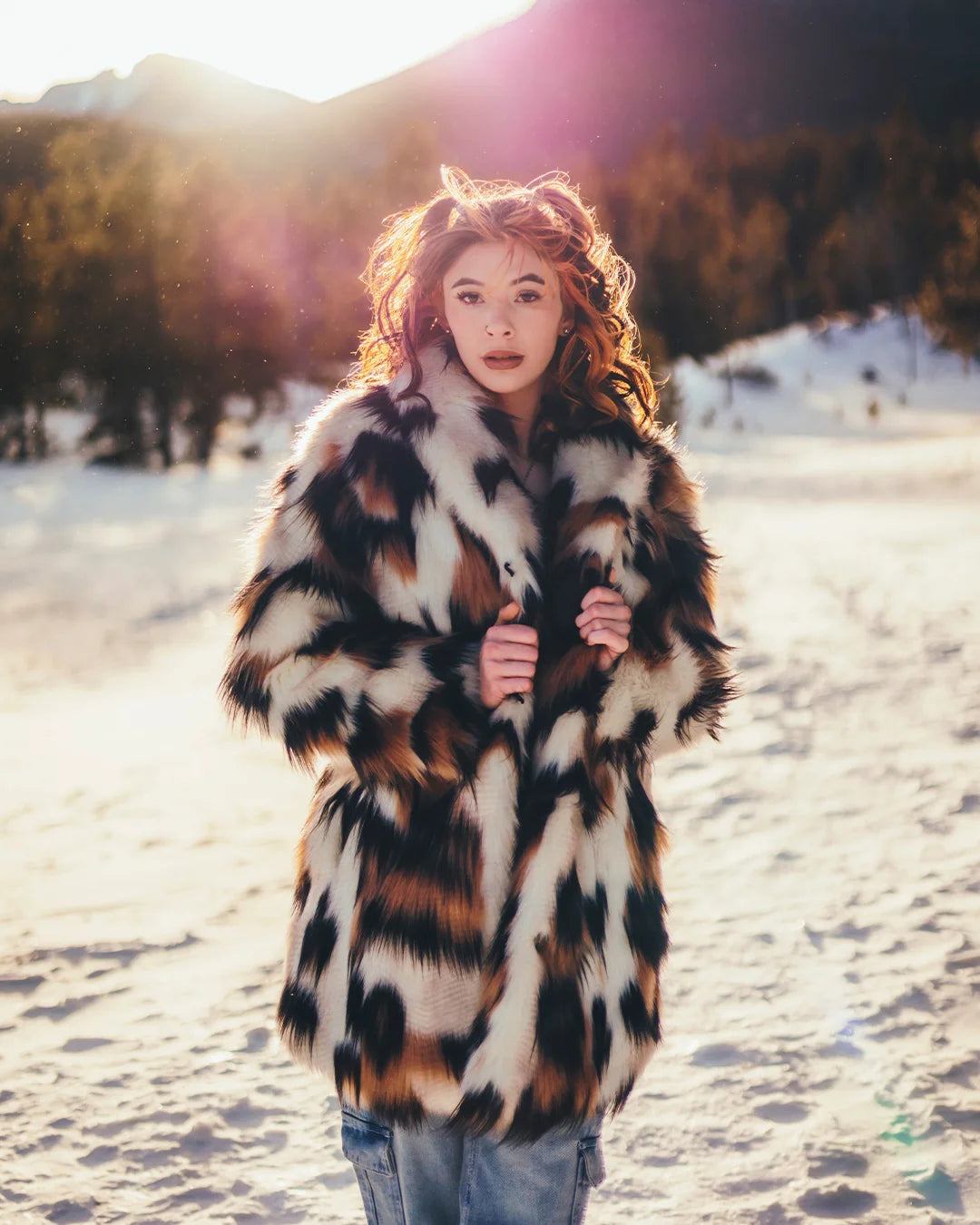 Woman wearing a patterned patchwork style plush faux fur coat standing in a snowy landscape with mountains in the background. SpiritHoods Collector Edition Manx Cat brown, black, and white fake fur jacket with an oversized collar