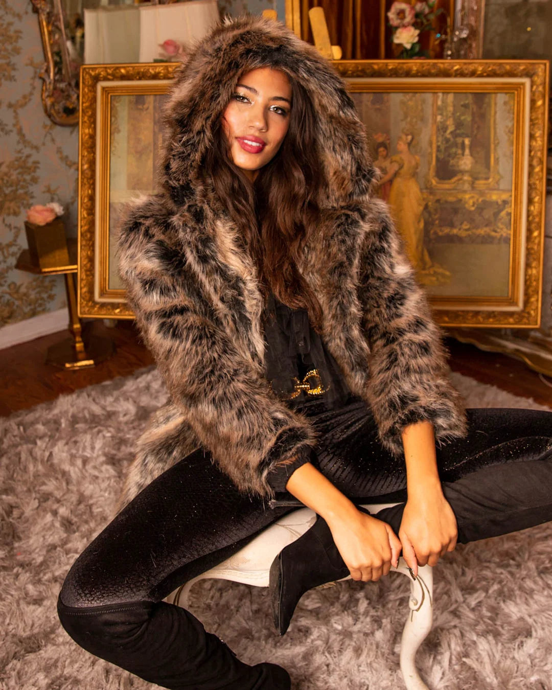 Smiling dark haired female model leaning against a painting while wearing hooded grey wolf fake fur coat.