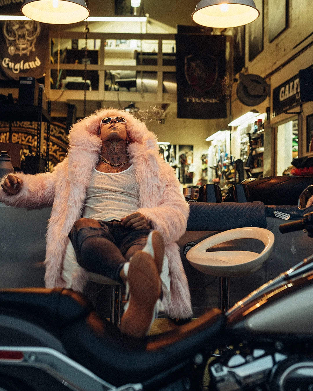 A Man sitting in a mechanics shop, with his legs up on a motorcycle and his head tilted back, wearing spirithoods Classic Hooded Pink Faux Fur Coat in Flamingo Wolf