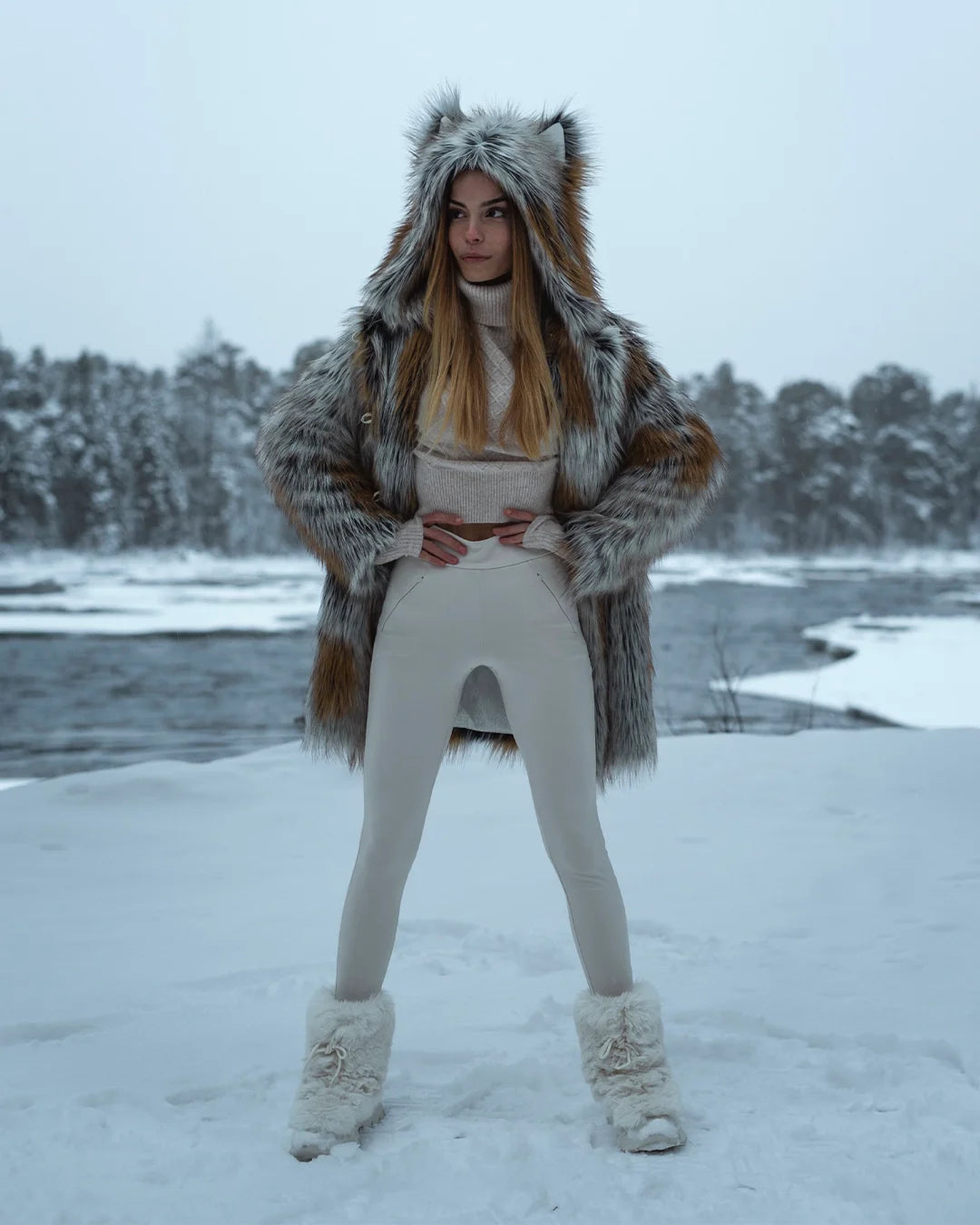 A Woman standing in Wonder Woman pose, looking out to the right, holding her hands at her waist as she stares into a winter wonderland, while wearing her plush SpiritHoods Arctic Fox 100% Vegan Fake Fur Coat  in a snowy landscape