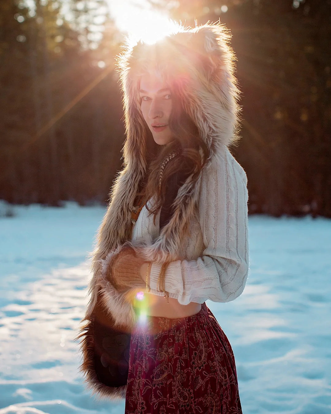 SpiritHoods Original Red Fox Faux Fur Hood on a model in nature, posing on a wooden fence, with fresh snow behind her, and shes snuggled up with her Red Fox SpiritHood, Hood, looking at the camera.