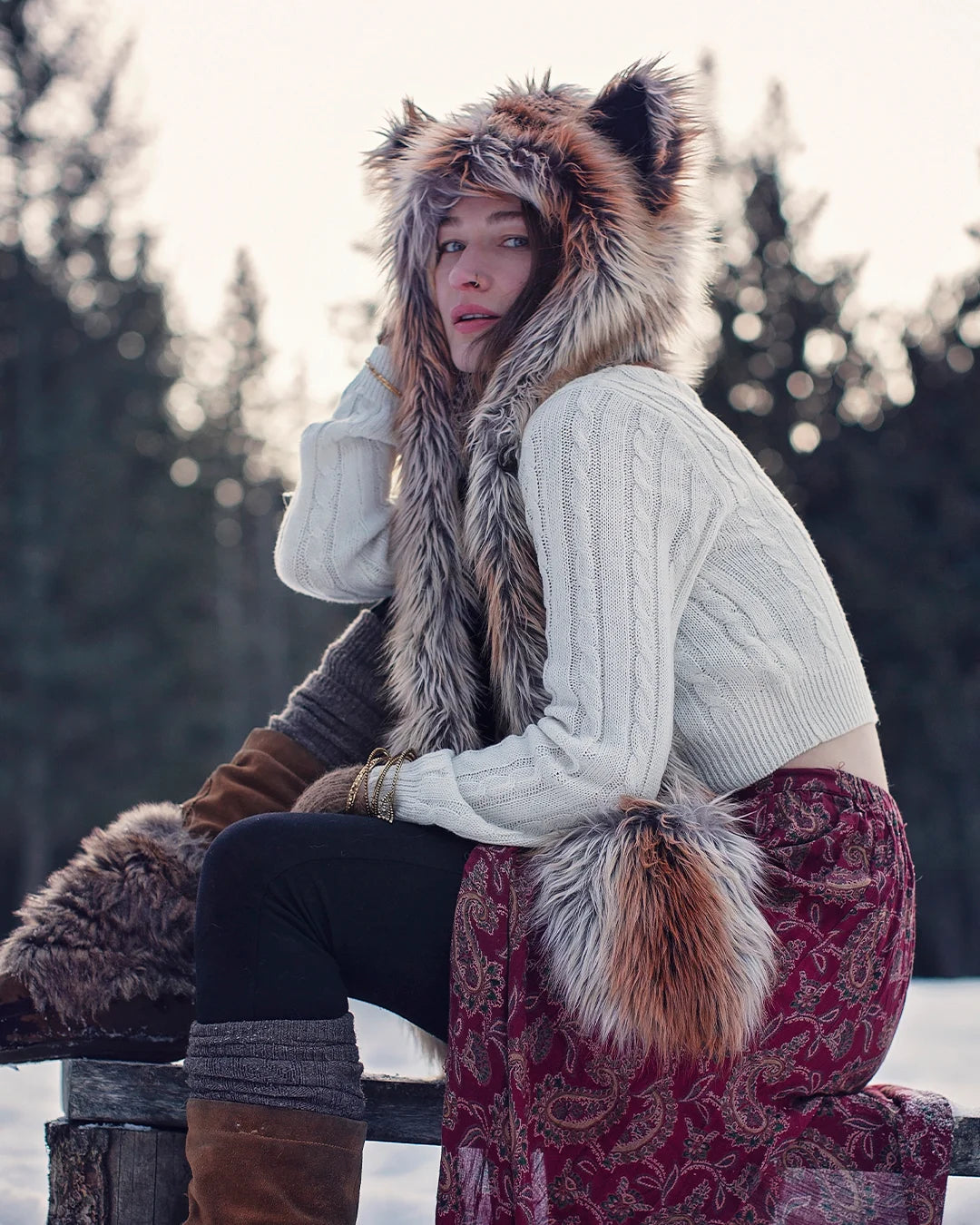 SpiritHoods Original Red Fox Faux Fur Hood on a model in nature, posing on a wooden fence, with fresh snow behind her, and shes snuggled up with her Red Fox SpiritHood, Hood, looking at the camera.