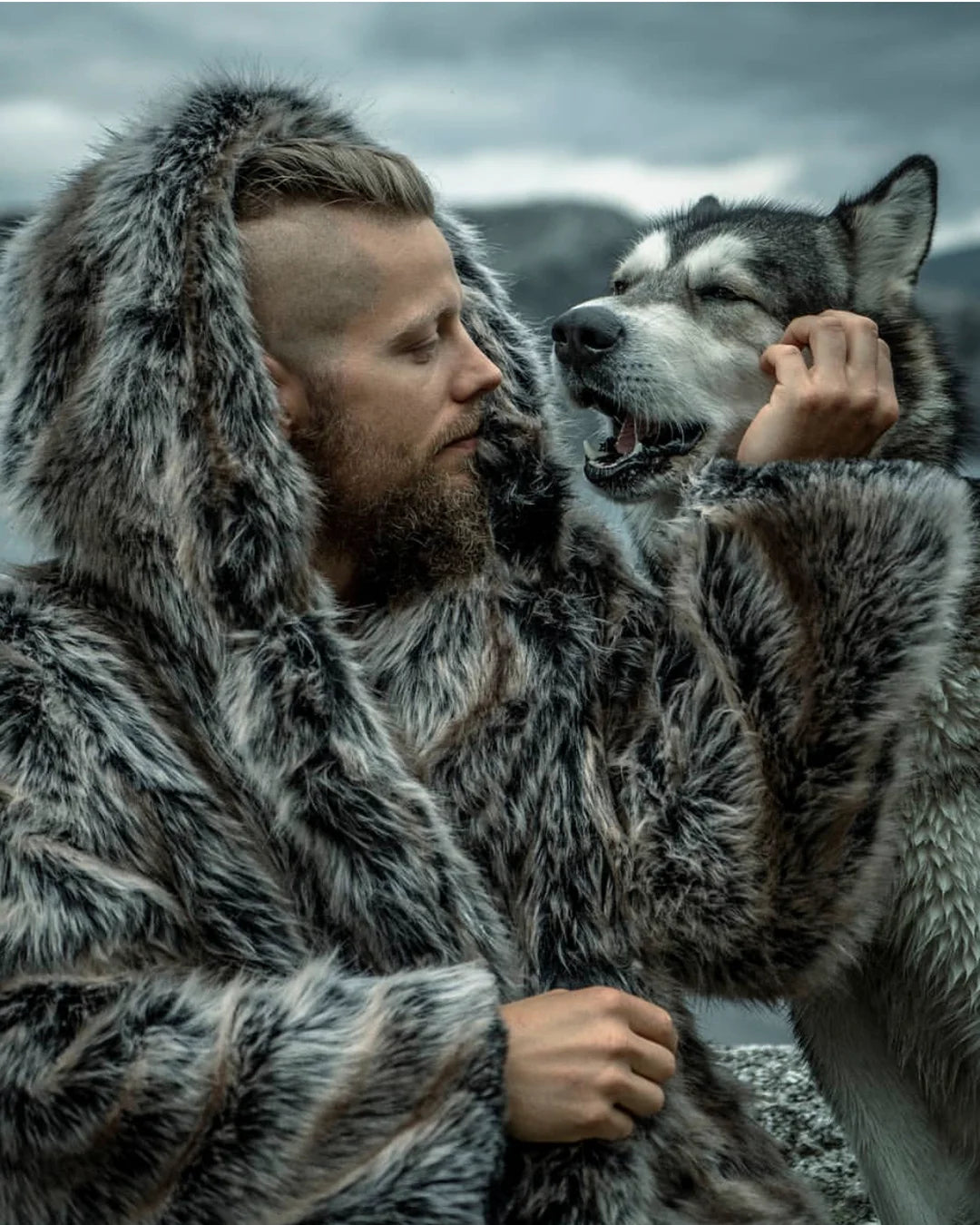A Man wearing SpiritHoods Grey Wolf Hooded Faux Fur Coat in nature in the middle of winter and snow, walking up the mountains with the plush oversized hood on.