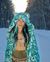 Model in the middle of the forest, with snow falling, wearing the SpiritHoods Aqua Cat Classic Collector Edition Faux Fur Coat, as she walks towards the camera 