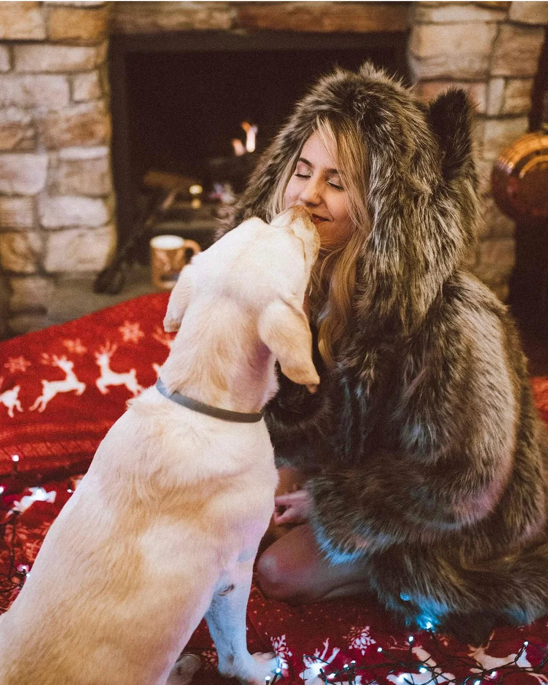 Woman in a Grey Wolf Classic Fake Fur Jacket by SpiritHoods sitting on a red christmas blanket with a dog, fireplace in the background