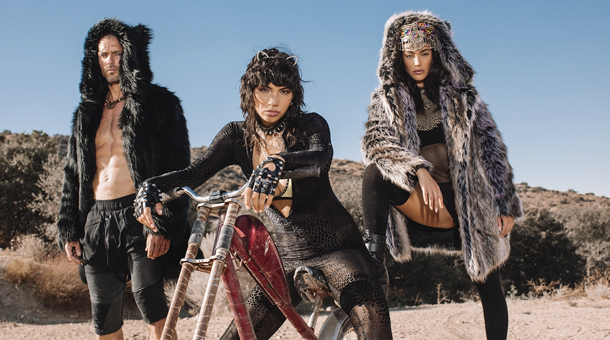 Three people in fur-lined hooded coats in a desert setting with a bicycle.