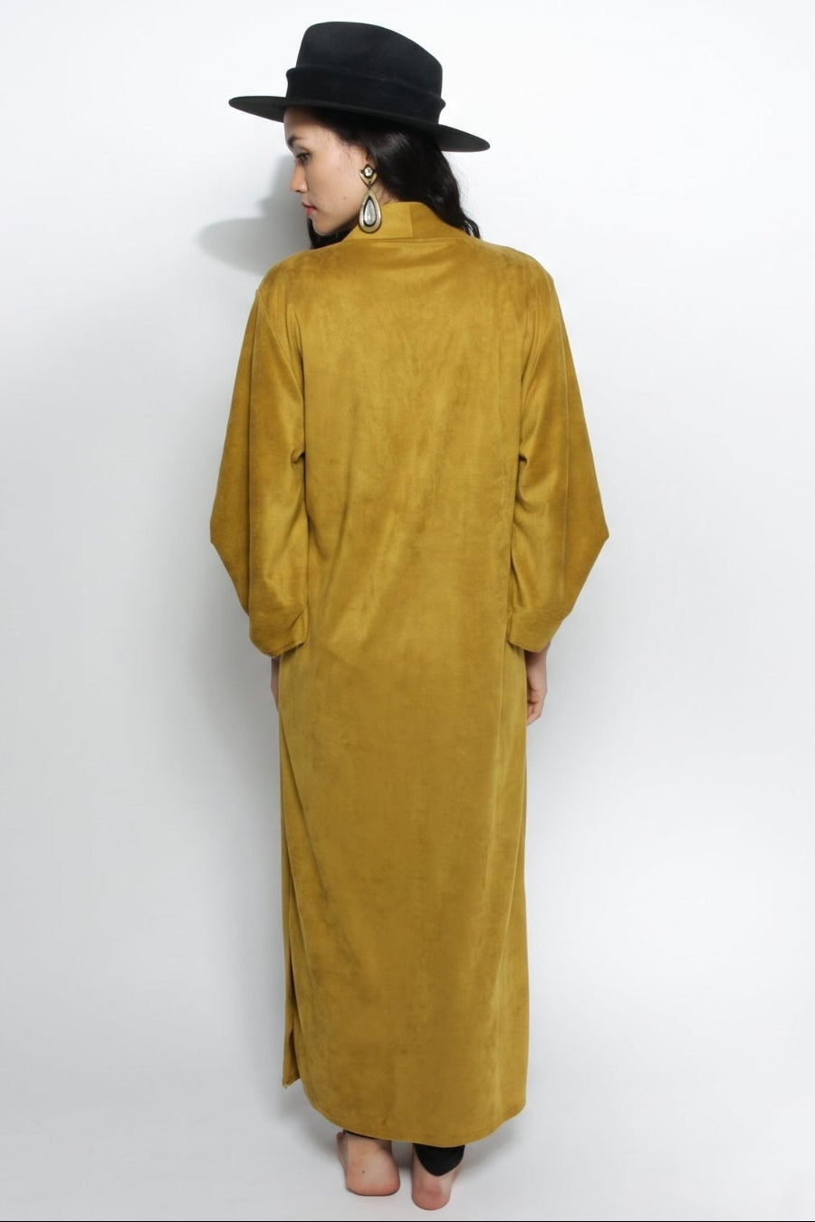 A woman stands with her back to the camera wearing a long, wide-sleeved mustard yellow dress with a high collar and a black wide-brim hat against a white studio background.