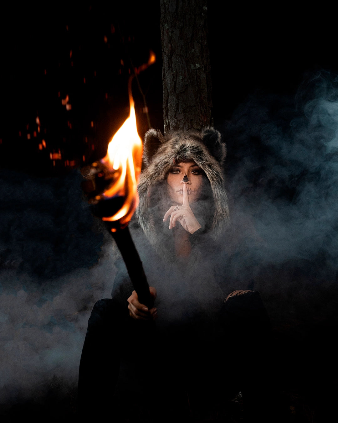 A Women wearing SpiritHoods Dire Wolf Faux Fur Shawl in the woods holding a stick that is on fire, as smoke comes up behind her. She has painted her nose black and 'stitched' her lips - easy halloween costume ideas for women