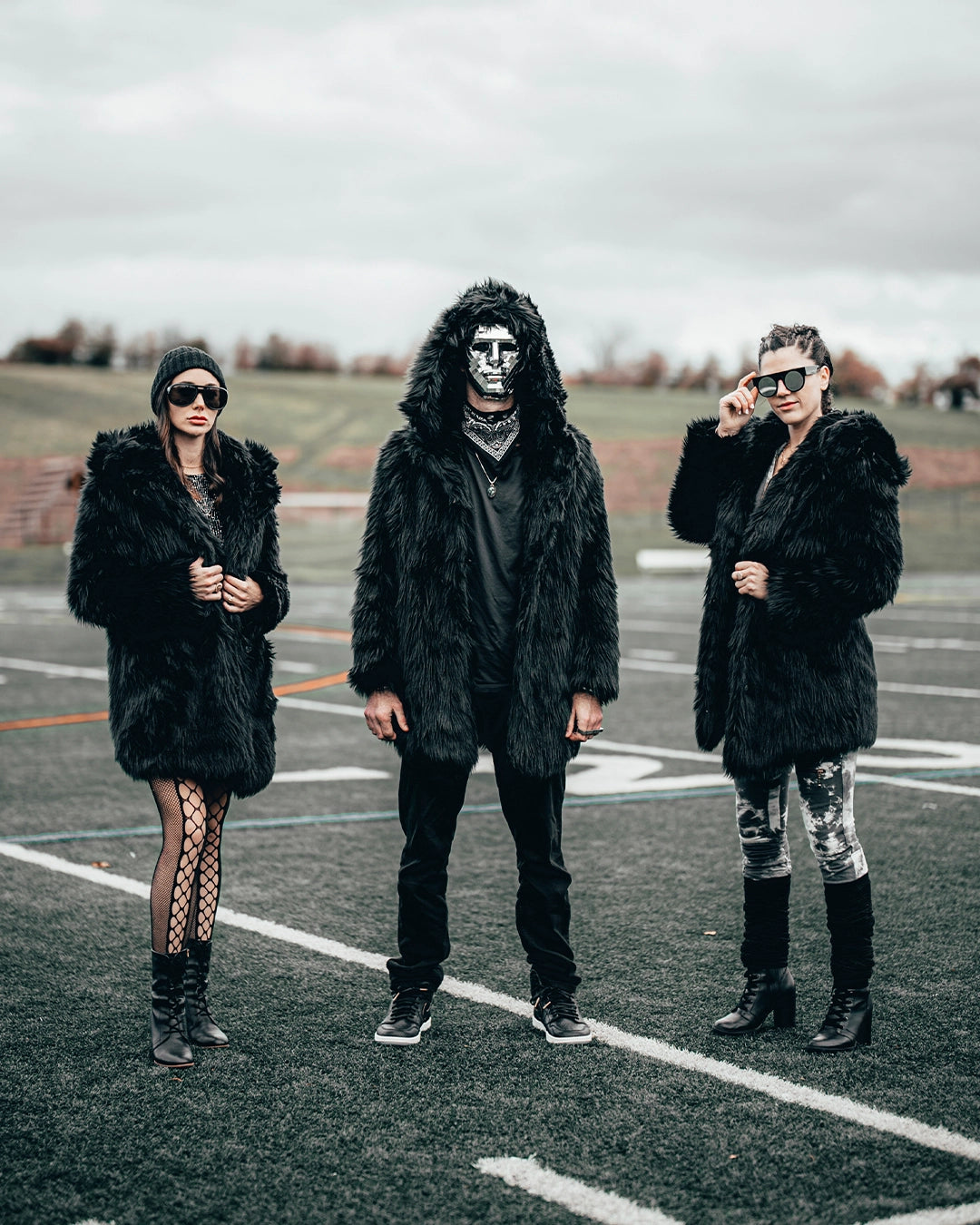 SpiritHoods Black Wolf Hooded Faux Fur Coat with a man standing on a football field wearing a halloween disco inspired mask with the black wolf hood on staring at the camera, two females are next to him wearing black wolf coats with the hoods off, looking at the camera 