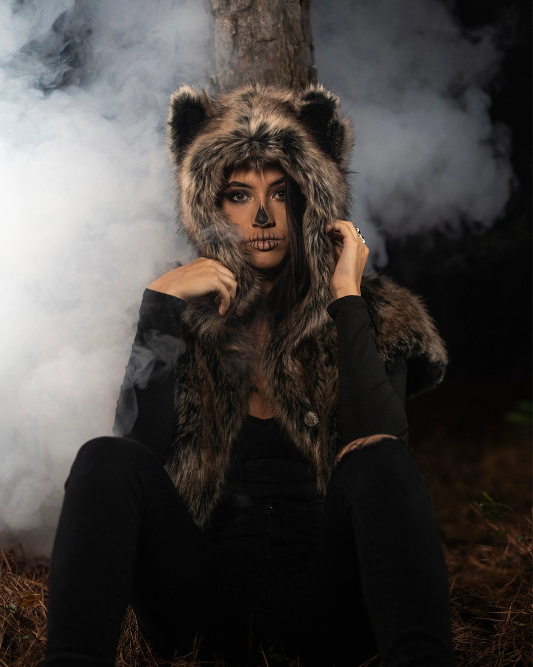 A Person with easy skull make up, sitting against a tree, in a forest at night, with smoke coming up behind her, wearing spirithoods best selling dire wolf fake fur shawl with wolf ears. Easy Halloween Outfits, Halloween Costumes.