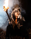 A Women wearing SpiritHoods Dire Wolf Faux Fur Shawl in the woods holding a stick that is on fire, as smoke comes up behind her. She has painted her nose black and 'stitched' her lips - easy halloween costume ideas for women