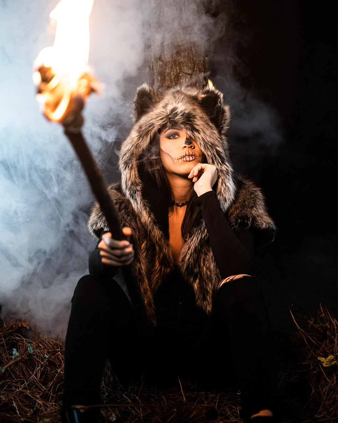 A Women wearing SpiritHoods Dire Wolf Faux Fur Shawl in the woods holding a stick that is on fire, as smoke comes up behind her. She has painted her nose black and 'stitched' her lips - easy halloween costume ideas for women
