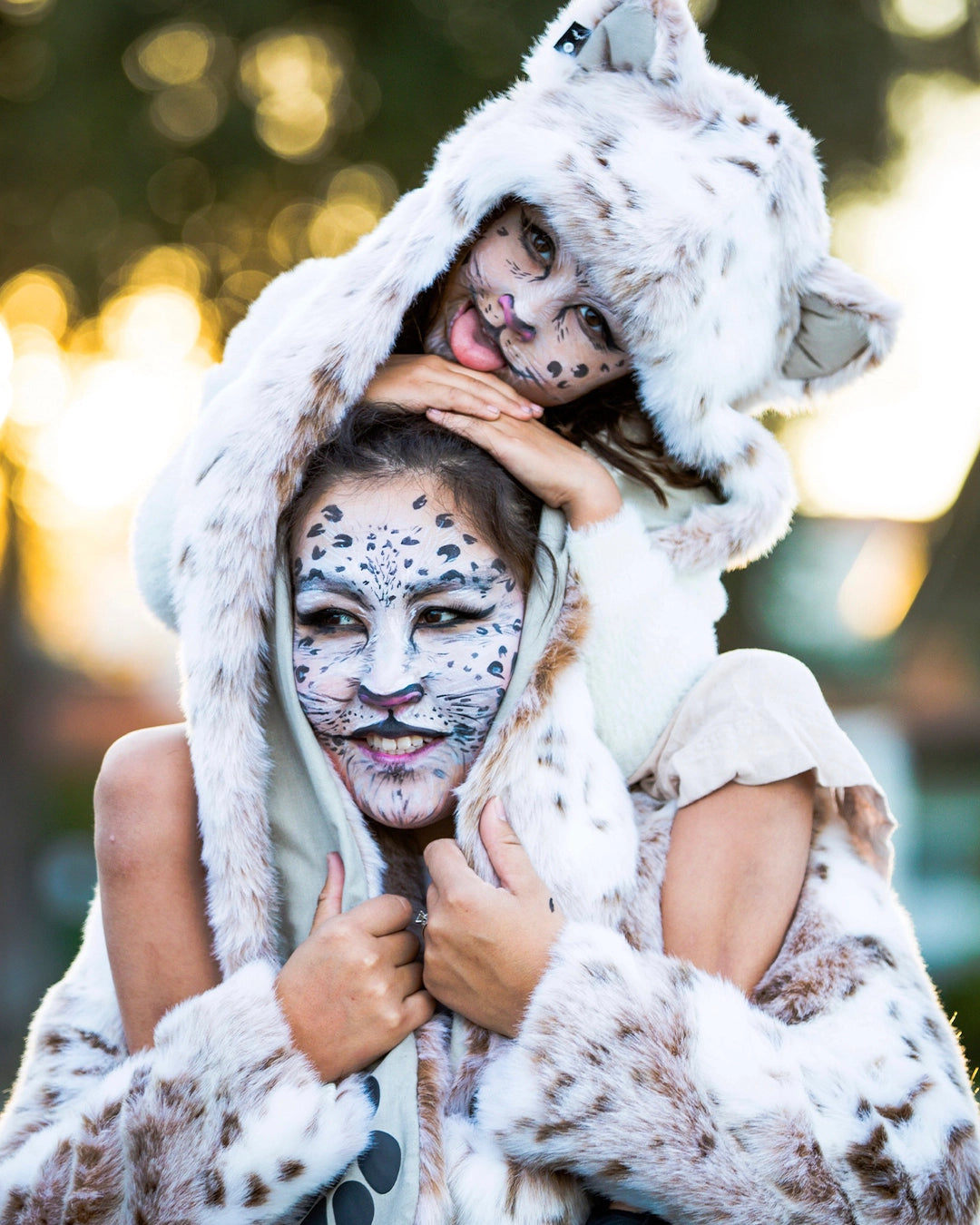 Two people in snow leopard-themed costumes with face paint against a blurred natural background. Wearing SpiritHoods Siberian Snow Leopard Fake Fur Hood with Paws and Ears, and Siberian Snow Leopard Fake Fur Jacket. Easy Halloween Costumes. Animal-themed halloween outfits. 