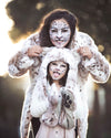 Two people in snow leopard-themed costumes with face paint against a blurred natural background. Wearing SpiritHoods Siberian Snow Leopard Fake Fur Hood with Paws and Ears, and Siberian Snow Leopard Fake Fur Jacket. Easy Halloween Costumes. Animal-themed halloween outfits. 