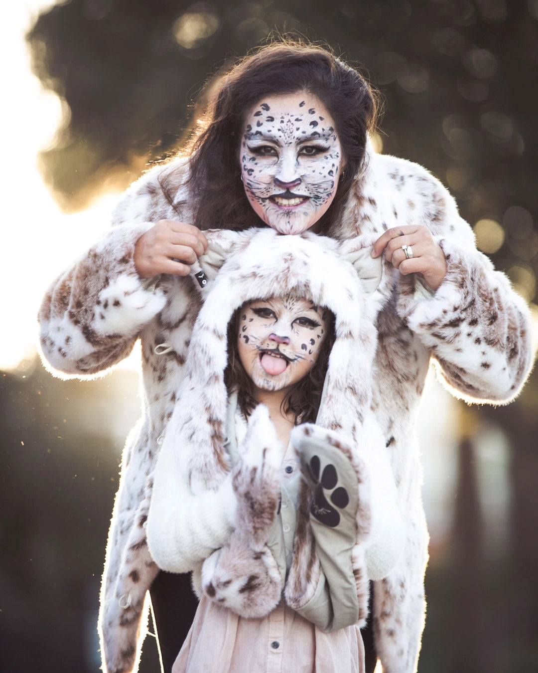 Two people in snow leopard-themed costumes with face paint against a blurred natural background. Wearing SpiritHoods Siberian Snow Leopard Fake Fur Hood with Paws and Ears, and Siberian Snow Leopard Fake Fur Jacket. Easy Halloween Costumes. Animal-themed halloween outfits. 