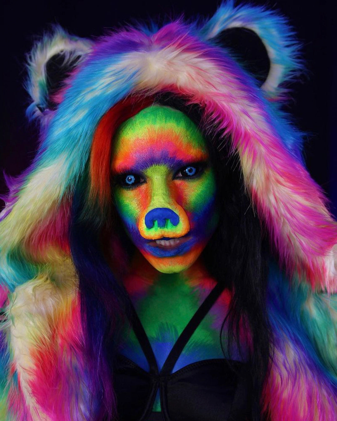 Woman with colorful rainbow tie-dye makeup with a bear nose and SpiritHoods uv-reactive Classic Rainbow Bear Faux Fur Coat with the hood up featuring bear ears, against a black background. Halloween Costume Ideas. Animal-themed costumes.