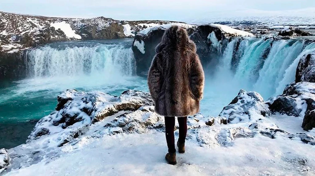 Person in a faux fur coat standing in front of a snowy waterfall.