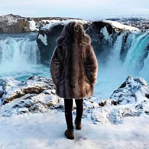 Person wearing a fur coat standing in front of a waterfall in a snowy landscape.