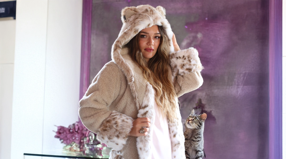 Woman wearing a fluffy hooded coat with a cat on a table next to her.