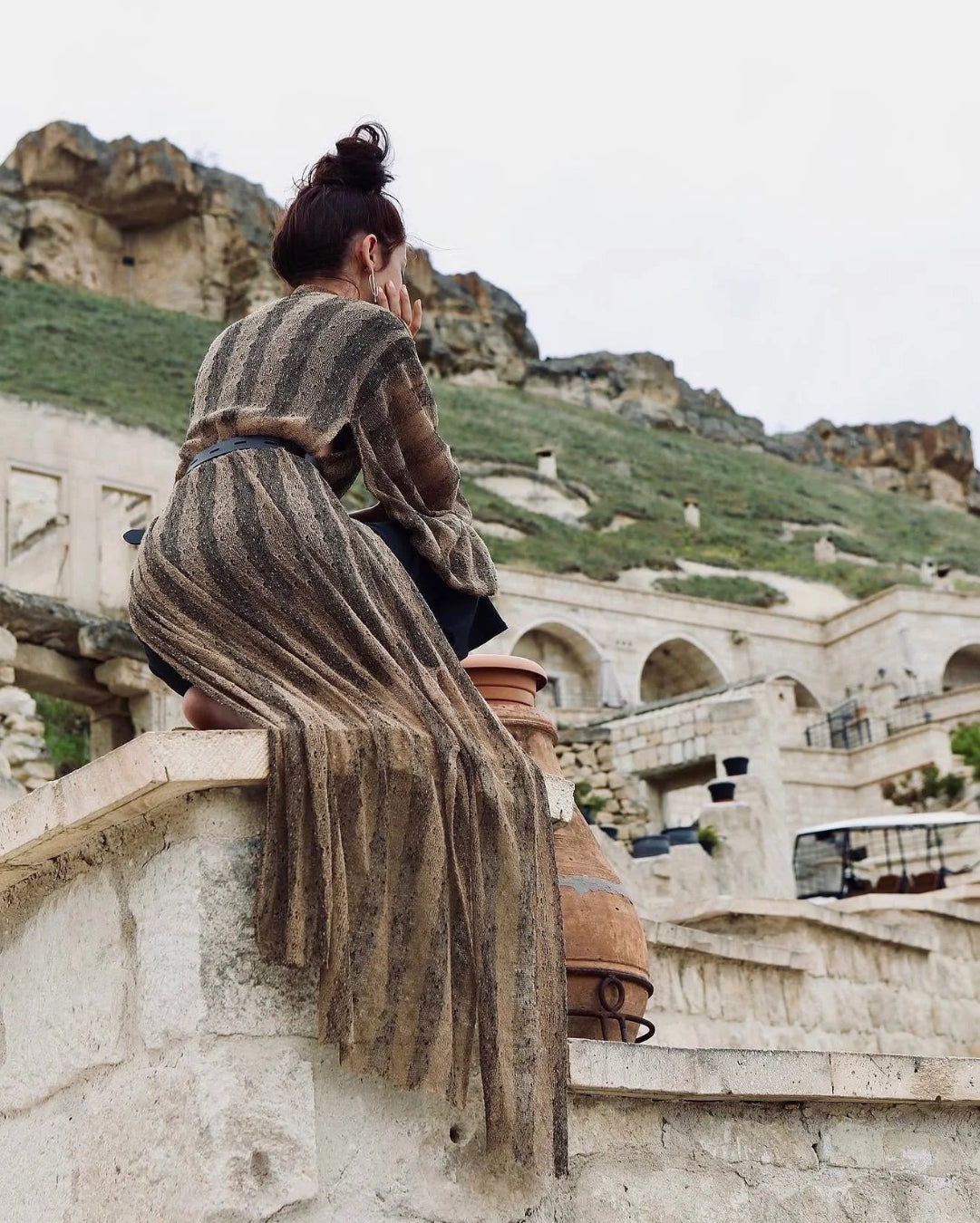 A woman wearing a brown and beige striped fringed shawl sits on a stone wall overlooking ancient cave dwellings and a grassy cliffside under an overcast sky.