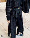 A person wearing a long black textured open-front coat with wide sleeves, a black belt, black top and leggings, and dark flats, standing on a stone walkway.