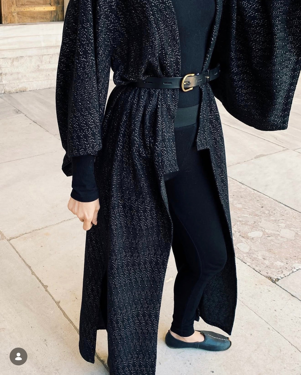 A person wearing a long black textured open-front coat with wide sleeves, a black belt, black top and leggings, and dark flats, standing on a stone walkway.