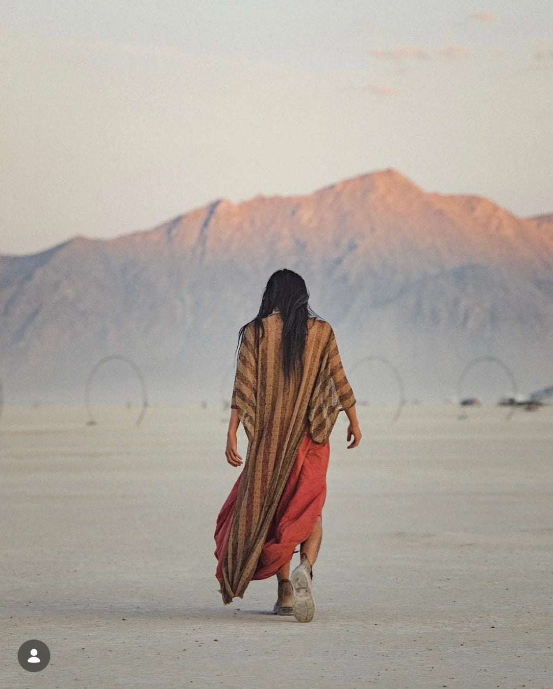 A woman seen from behind walking across a vast desert plain toward distant mountains, wearing a long brown striped wrap and a rust-red dress or skirt.