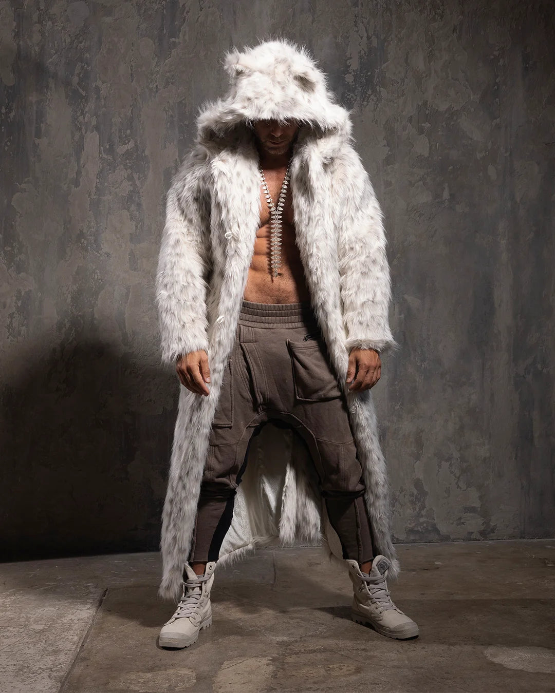 A Man wearing a long white fake fur coat with snow leopard print with a hood against a gray textured wall.