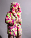 A man wearing a vibrant, multicolored neon fur hooded jacket with pink, green, and purple tones, standing against a plain gray studio background.