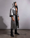 A man modeling a long, hooded robe with a metallic silver and black patterned exterior and solid black lining, worn open over a bare torso, black pants, and black boots, standing against a plain gray studio backdrop.