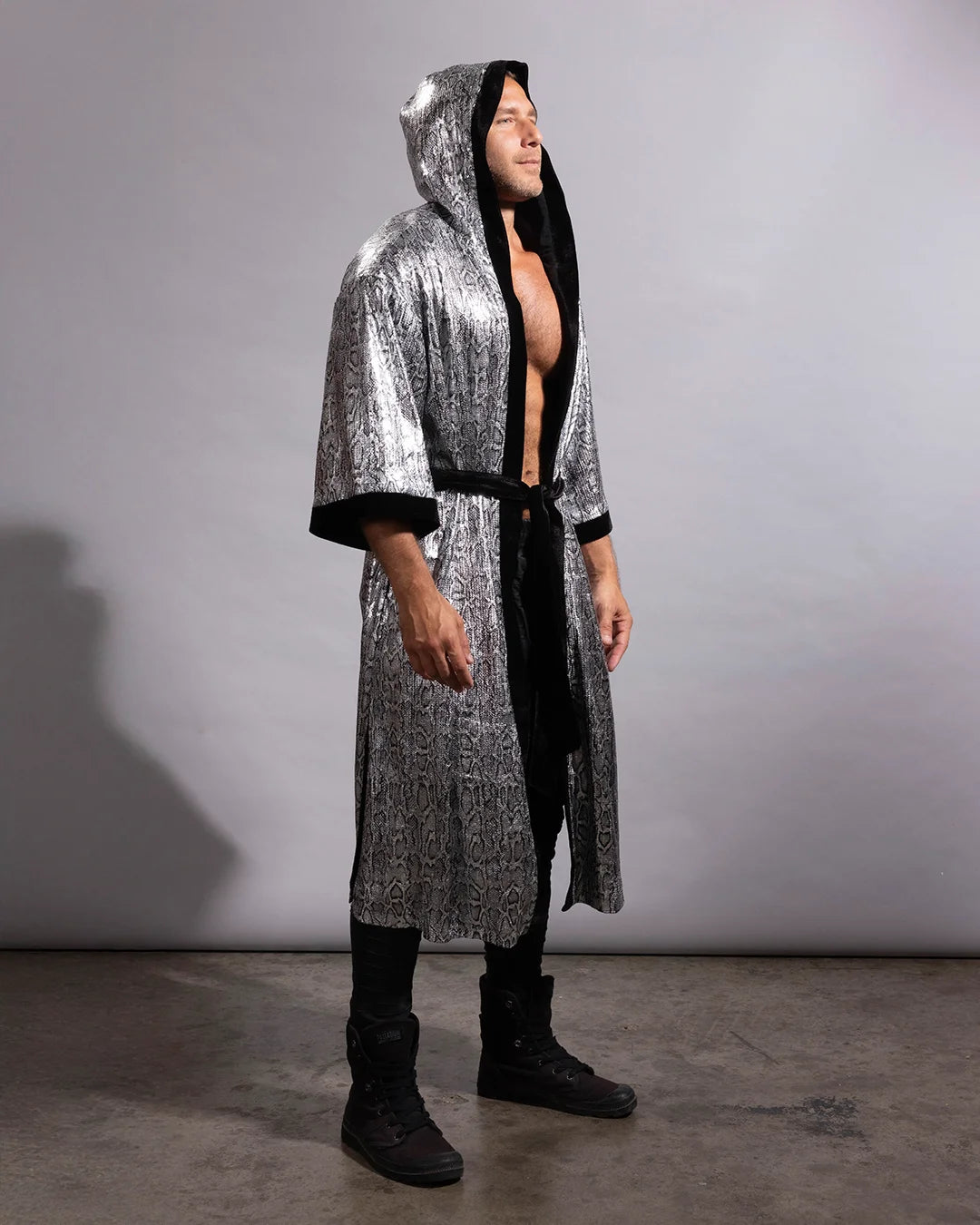 A man modeling a long, hooded robe with a metallic silver and black patterned exterior and solid black lining, worn open over a bare torso, black pants, and black boots, standing against a plain gray studio backdrop.
