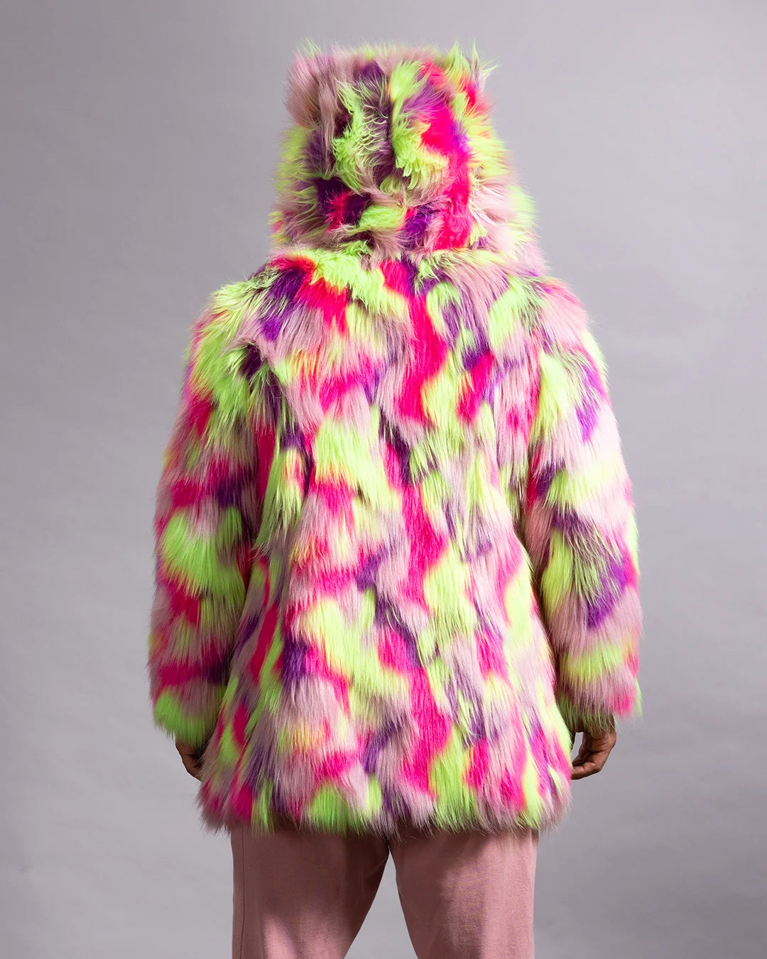 A person shown from the back wearing a hooded faux fur jacket with a vibrant neon tie-dye pattern in pink, lime green, and purple, paired with dusty pink pants, against a neutral gray studio background.