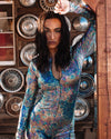 A woman posing in a metallic silver rave costume bodysuit with a holographic snakeskin pattern, standing in front of a backdrop of car hubcaps.