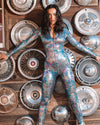 A woman posing in a Reflective rave outfit bodysuit with a holographic snakeskin pattern, standing in front of a backdrop of car hubcaps.