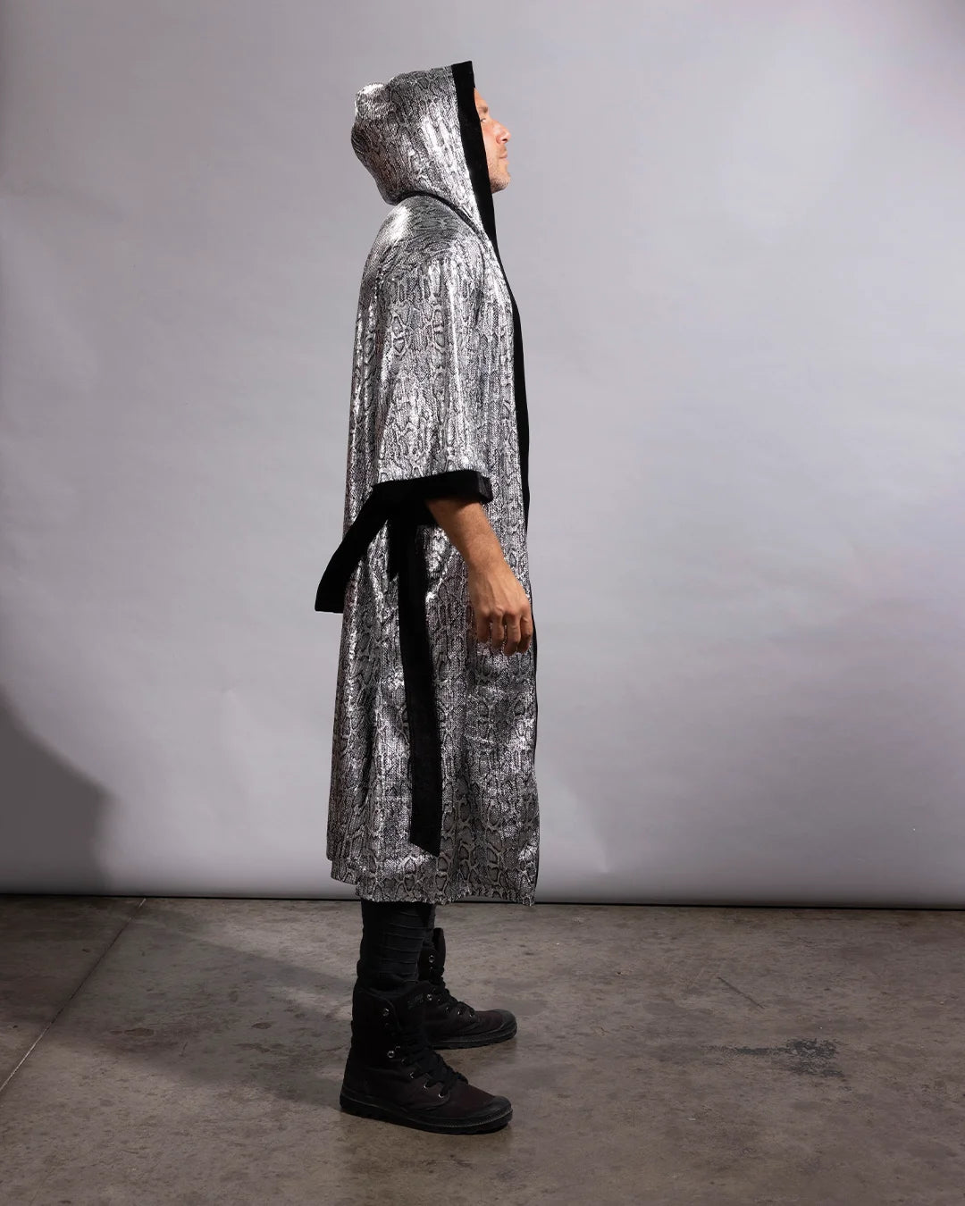 A person wearing a long silver sequin hooded robe with black trim and a black waist tie, standing in profile against a plain light gray studio backdrop.