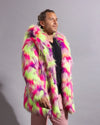 A man wearing a vibrant, multicolored neon fake fur jacket with pink, lime green, and purple patterns, paired with a black shirt and pink pants, standing against a plain gray studio background.