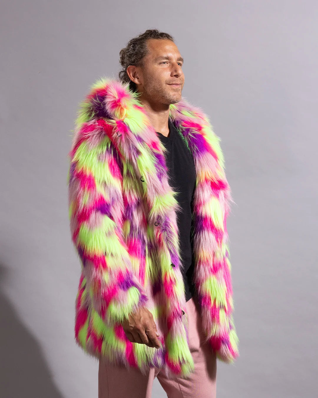 A man wearing a vibrant, multicolored neon fake fur jacket with pink, lime green, and purple patterns, paired with a black shirt and pink pants, standing against a plain gray studio background.