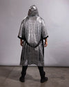 A person viewed from the back wearing a long, hooded silver robe with a black belt and black trim, standing against a plain light gray studio wall and concrete floor.