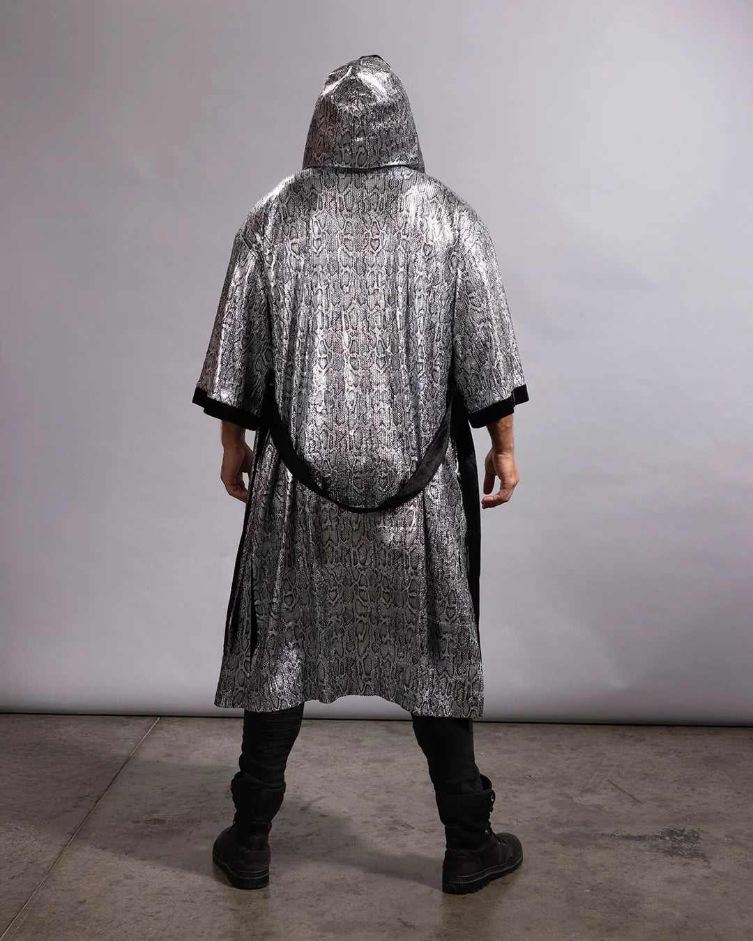 A person viewed from the back wearing a long, hooded silver robe with a black belt and black trim, standing against a plain light gray studio wall and concrete floor.