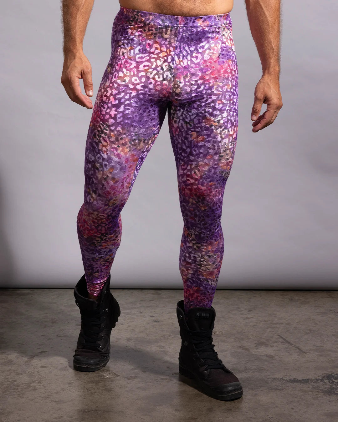 A model from the waist down wearing high-waisted rave meggings with a purple, pink, and orange leopard print pattern, paired with black boots against a gray studio background.