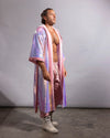 A man stands in profile wearing a sparkling iridescent sequin festival kimono over a matching pink outfit and white boots, photographed against a plain light gray studio backdrop.
